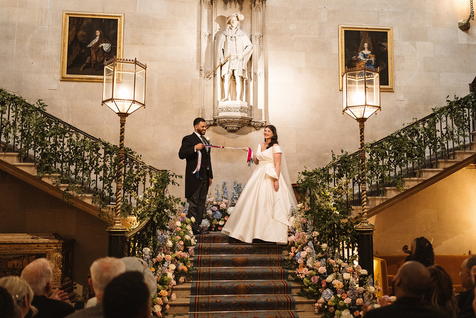 an american bride and groom standing on top of the stairs at Ashridge House, holding some colourful ribbons on both ends and smiling at the guests. The bride is in a beautiful white bridal gown and the groom is in a black tuxedo.