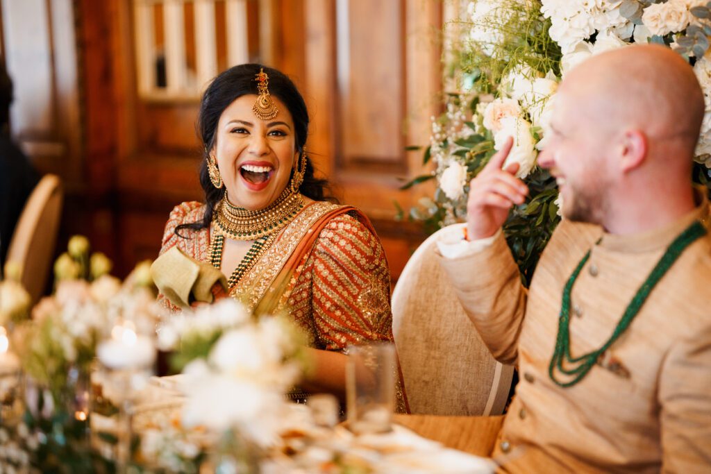 Indian bride laughing her heart out with her new polish husband. both the couple are in asian wedding attire. the bride is wearing a dark burn orange lehenga and the groom a beige sherwani. The bride has adorned beautiful Indian jewellery. They are both sitting on the sweetheart table during their wedding breakfast. The wedding venue is the hedsor house.