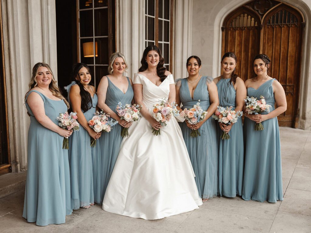 An American bride standing holding her bouquet with her bridesmaids who are all dressed in blue. There are 6 bridesmaids, 3 on either side of the bride. They are all holding bridesmaids' bouquets. this is taken in logia at Ashridge House