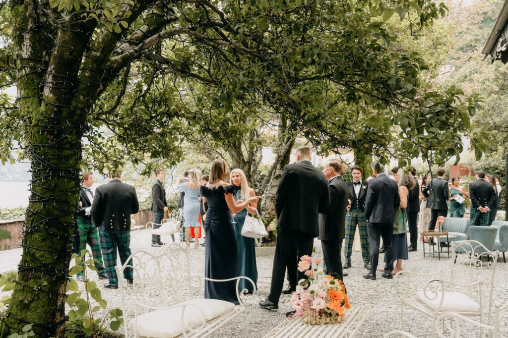 The image is of a canapes and drinks reception overlooking Lake Como. There are guests talking, eating canapes and holding drinks. It's a sunny day, but all guests seem to be under the shade of trees. 