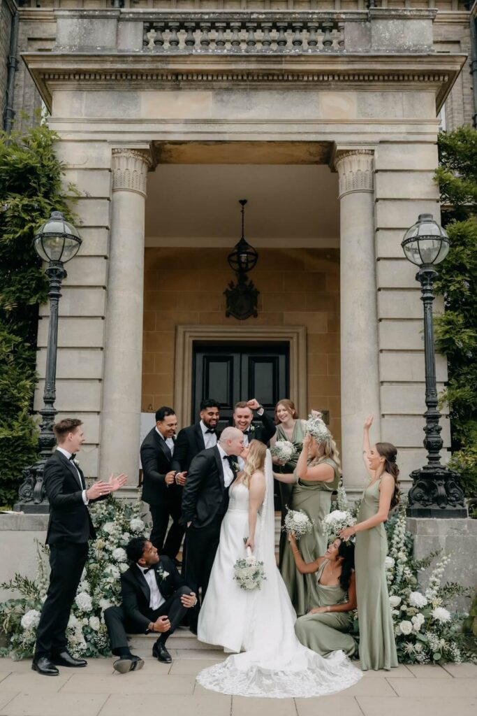 Wedding party celebrating with bride and groom on the steps of Hedsor House, Buckinghamshire, with sage green bridesmaid dresses and white hydrangea floral arrangements