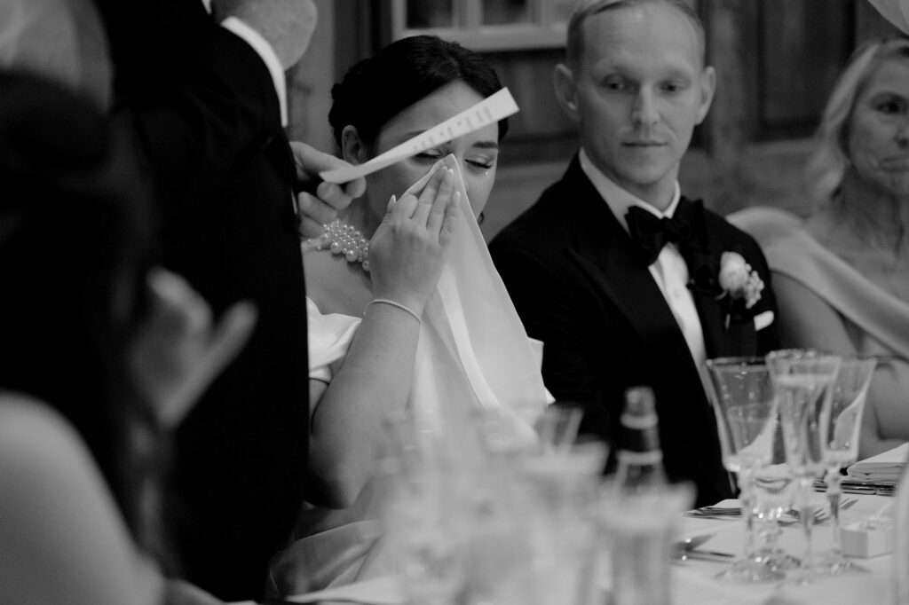 A beautiful British bride wiping her tears during her wedding breakfast, listening to her dad give his Father of the Bride speech, while an emotional groom looked on. The venue is Hedsor House.